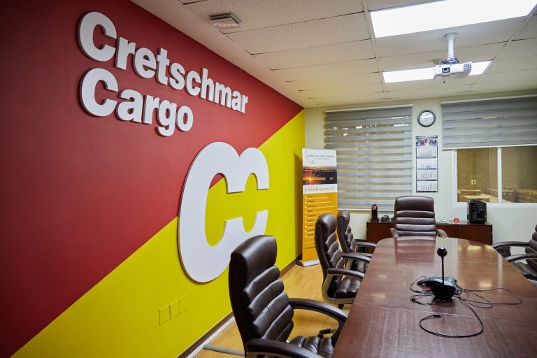 Besprechungsraum mit dem Logo von Cretschmar Cargo an der Wand, einem Holztisch und mehreren schwarzen Bürostühlen, sowie einem Projektor und einem Informationsbanner im Hintergrund.